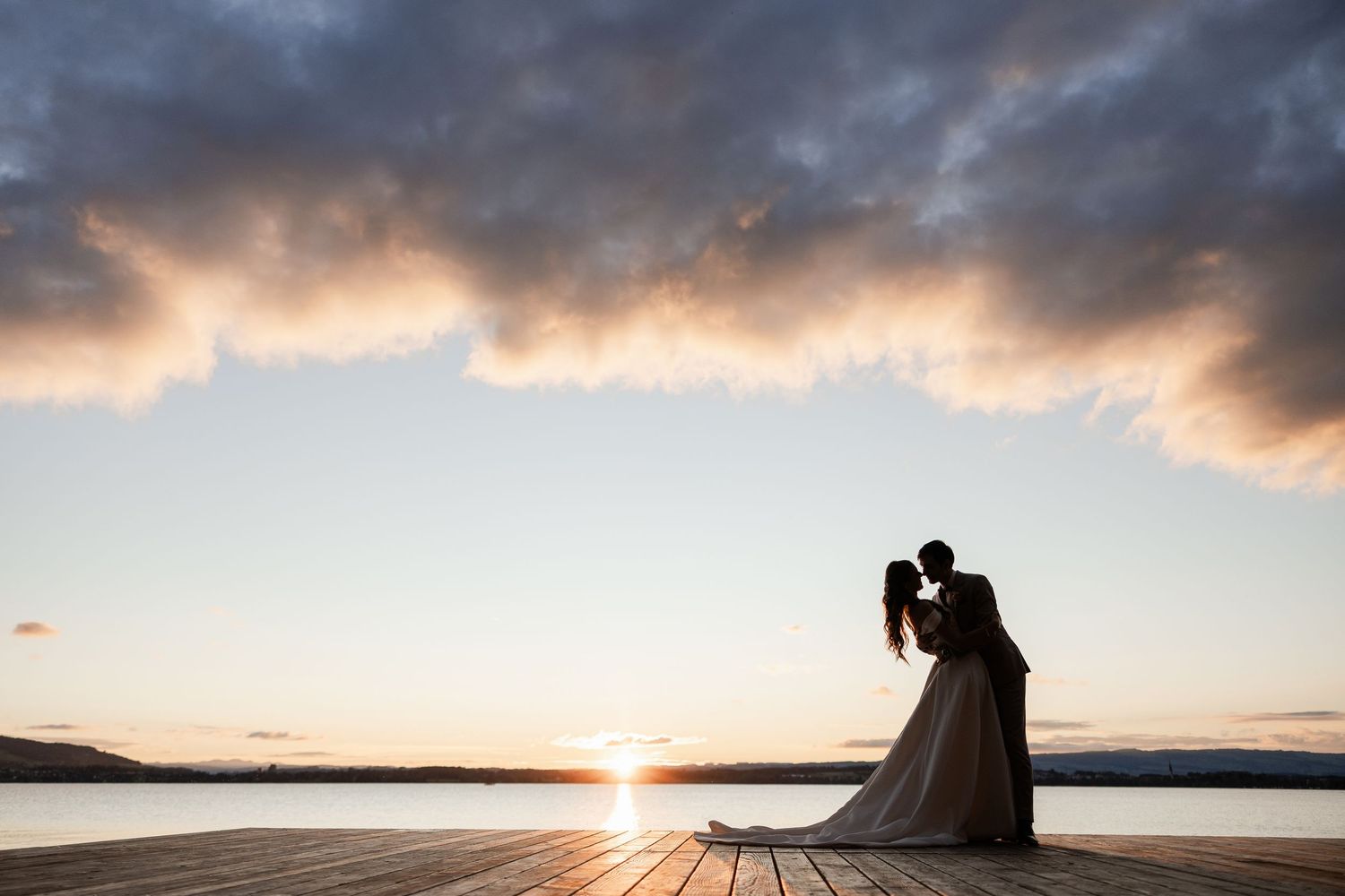 Silhouetted couple embraces on wooden pier during dramatic sunset over calm lake waters.