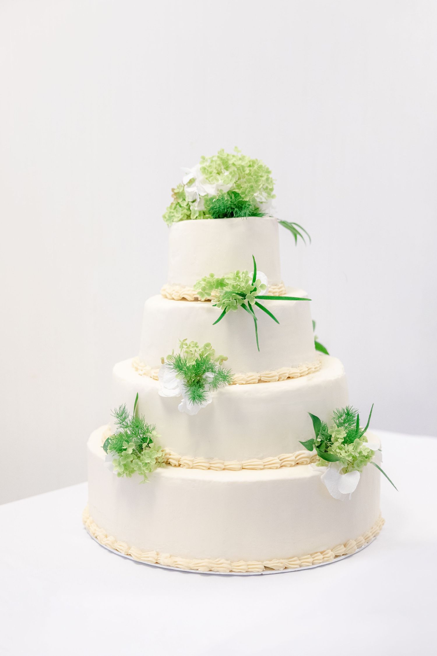 Four-tier white wedding cake decorated with delicate green florals and air plants on a white background.