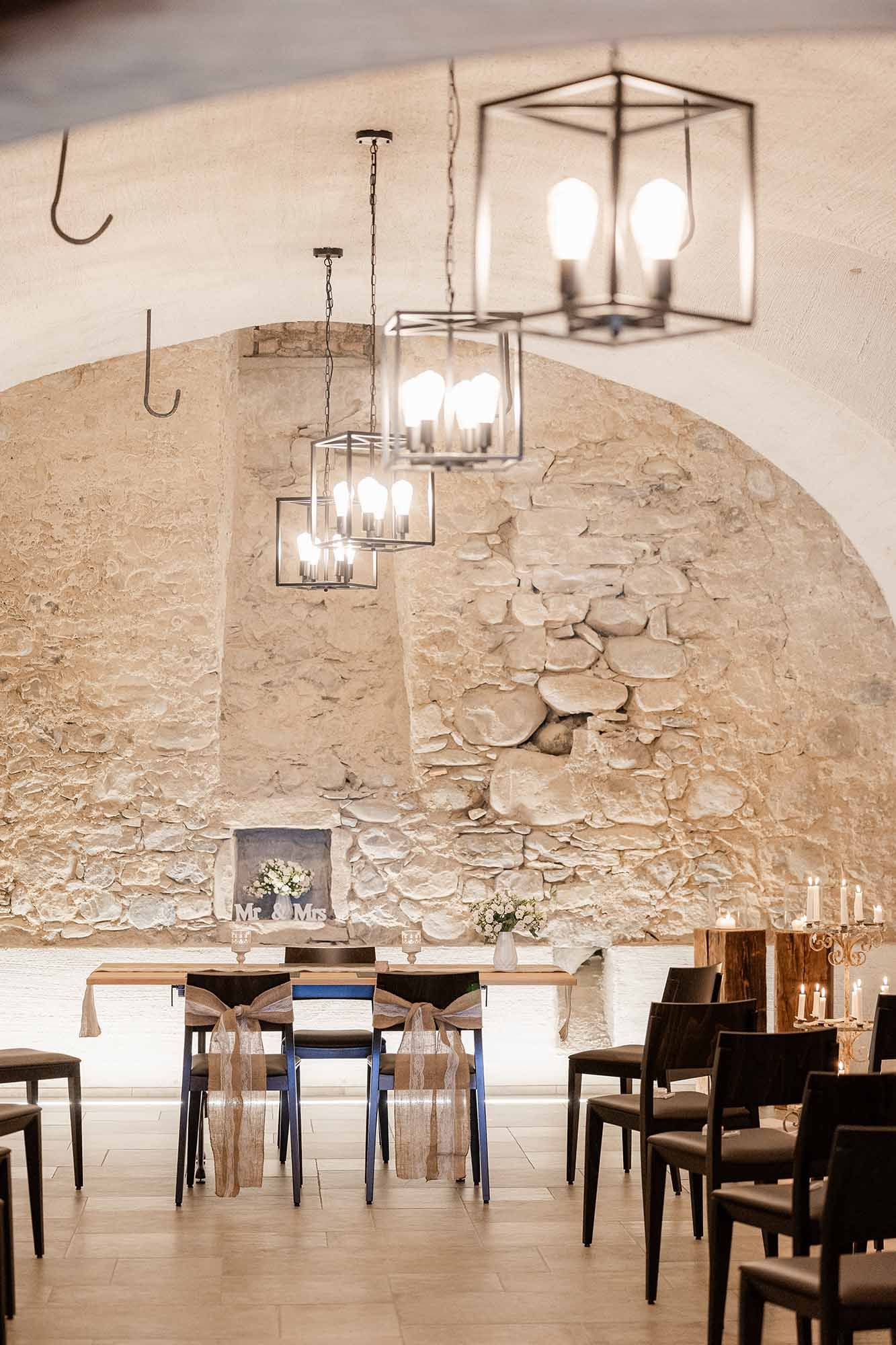 Rustic wedding reception venue with stone arch walls, hanging lantern chandeliers, and elegantly set dining tables with chairs.