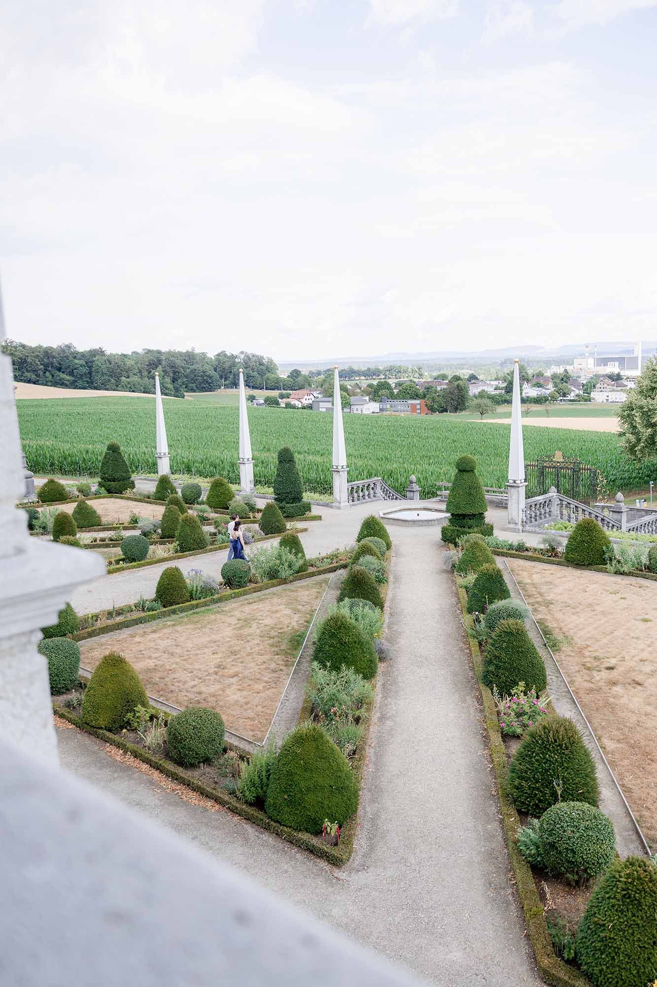 Formal European garden with manicured topiary hedges, cone-shaped shrubs, and tall obelisks overlooking green fields.