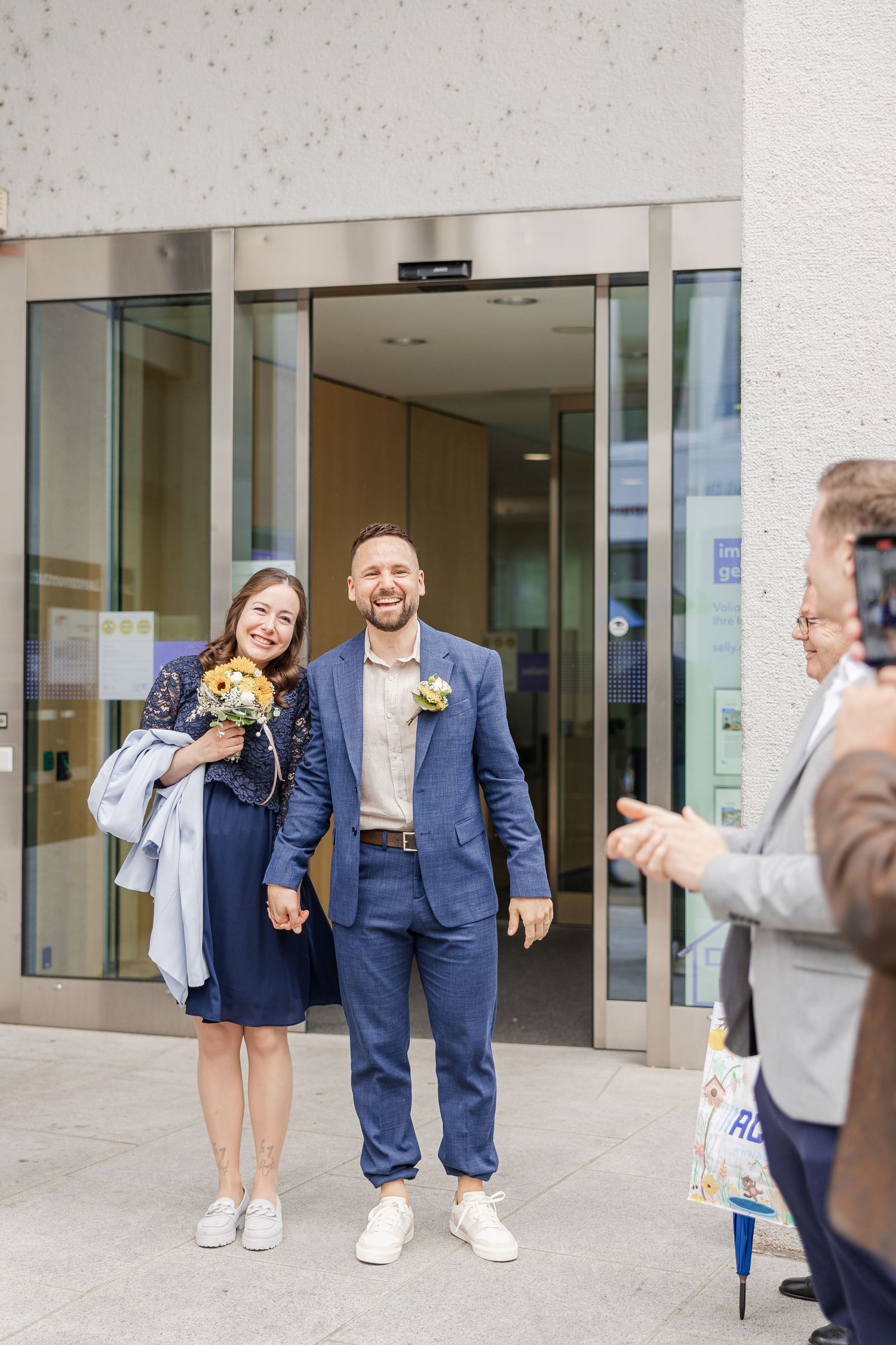 Happy couple exits building after civil wedding ceremony, groom in navy suit, bride in blue dress holding yellow flowers.