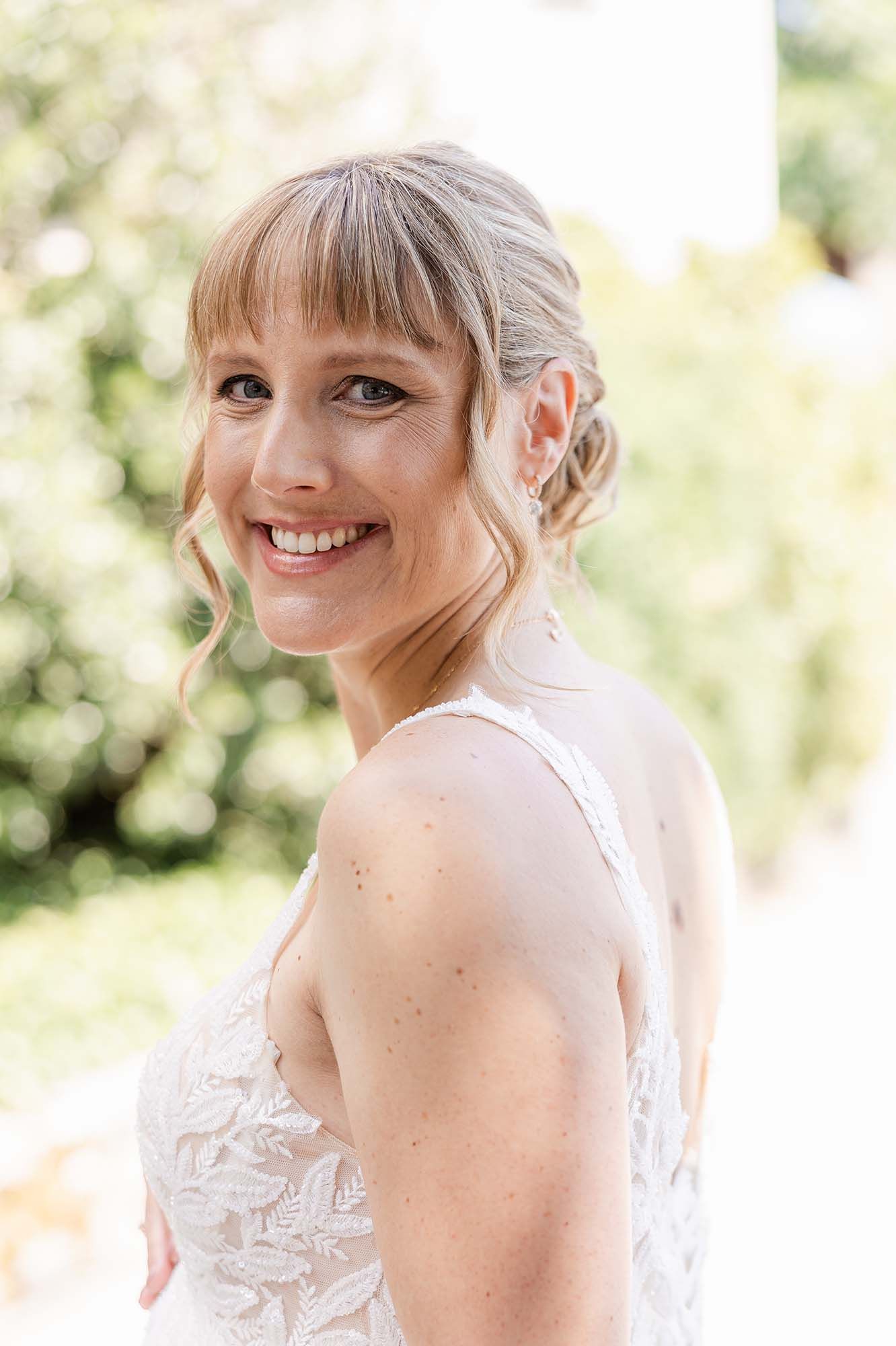 Smiling bride in lace wedding dress with bangs and updo hairstyle posing outdoors in a sunny garden setting.