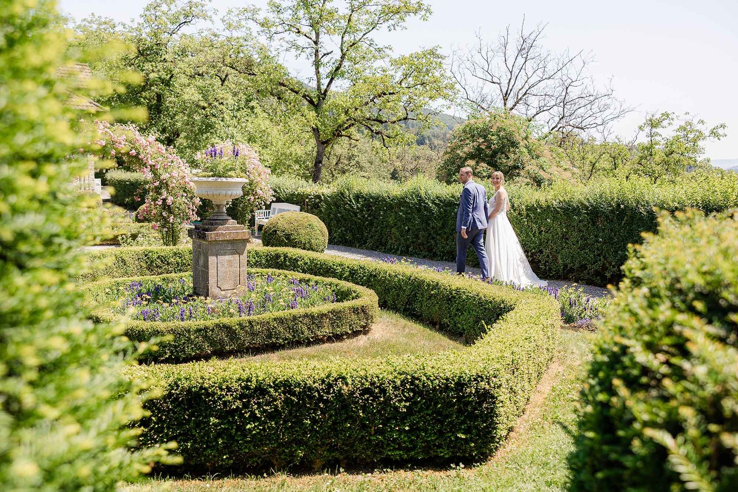 Bride and groom walking through elegant formal garden with manicured hedges and stone fountain on their wedding day.