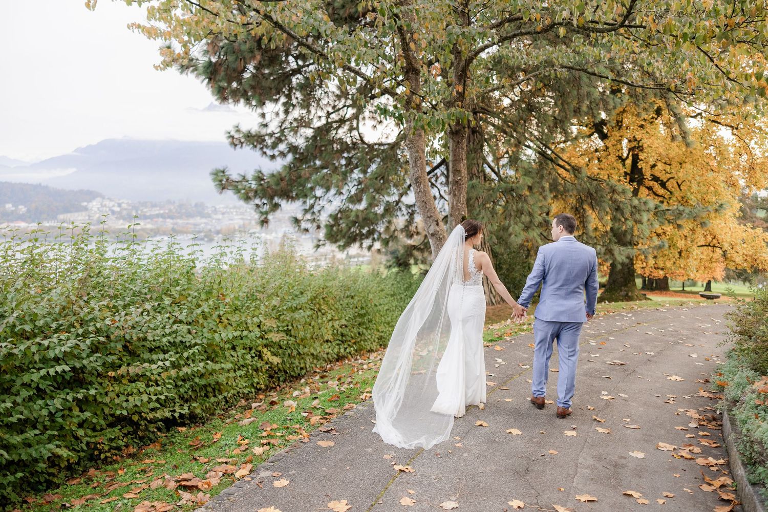 A couple walks along a lakeside path under autumn trees with mountains in the distance.