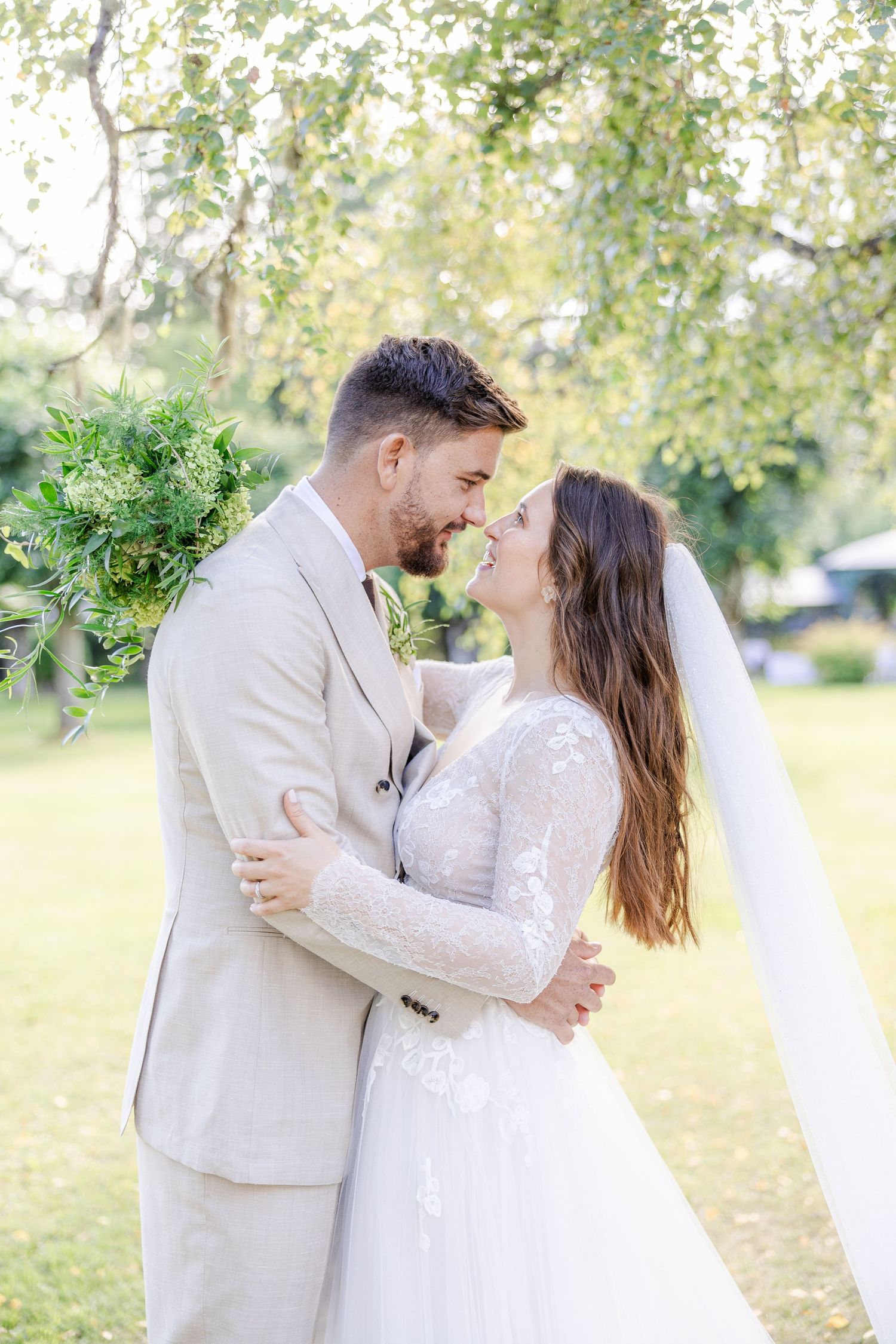 A couple in light-colored wedding attire embrace in a romantic outdoor setting with trees and natural light.