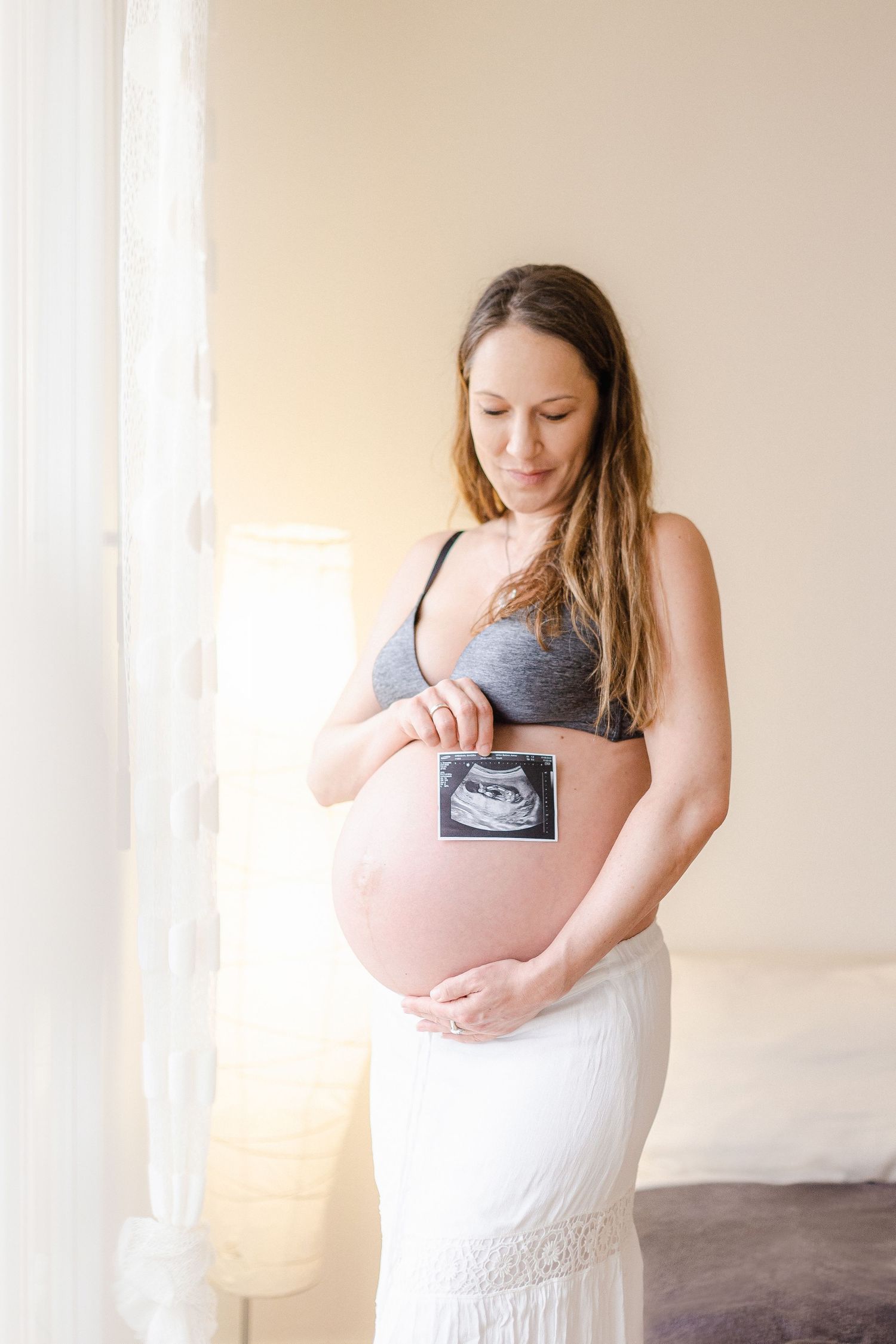 A pregnant person in a gray top and white skirt holds an ultrasound photo against their belly - Babybauch Fotograf Aargau
