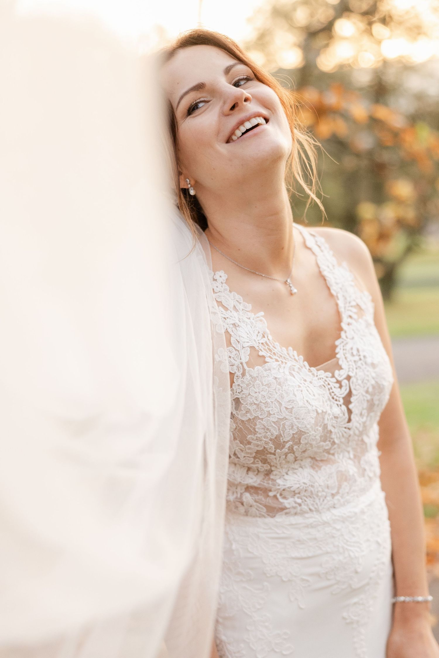 A bride in a delicate lace wedding dress laughs joyfully outdoors during a golden autumn photoshoot.
