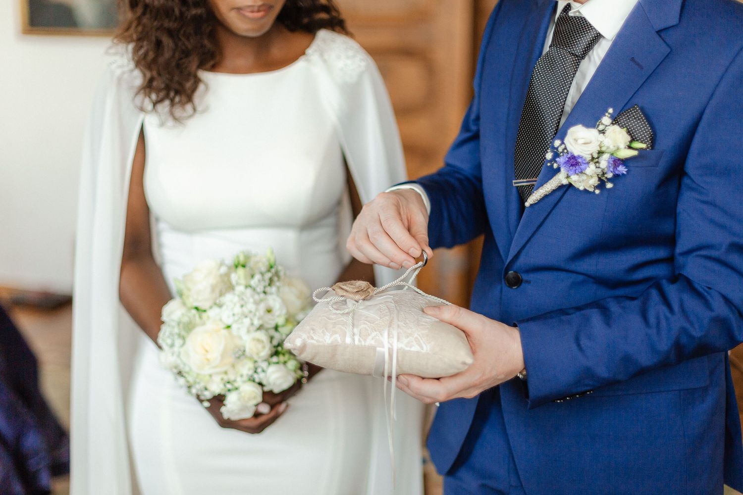 Closeup detail of wedding couple's hands with bouquet of white flowers, showing bride in white dress and groom in blue suit.