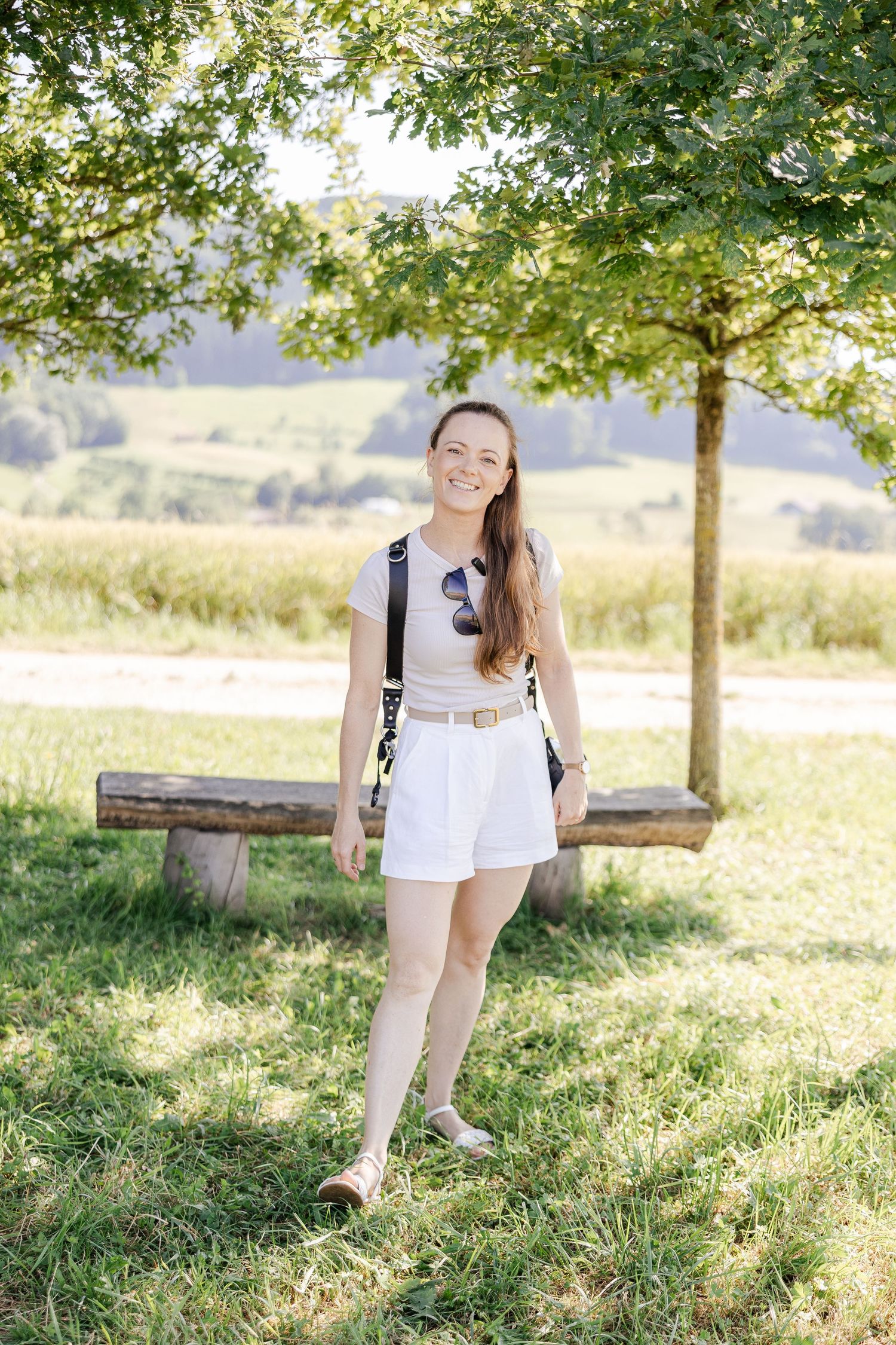 A person in white summer attire sits on a wooden log under green trees in a sunny meadow - Andrea Klimenko Hochzeitsfotograf Aargau