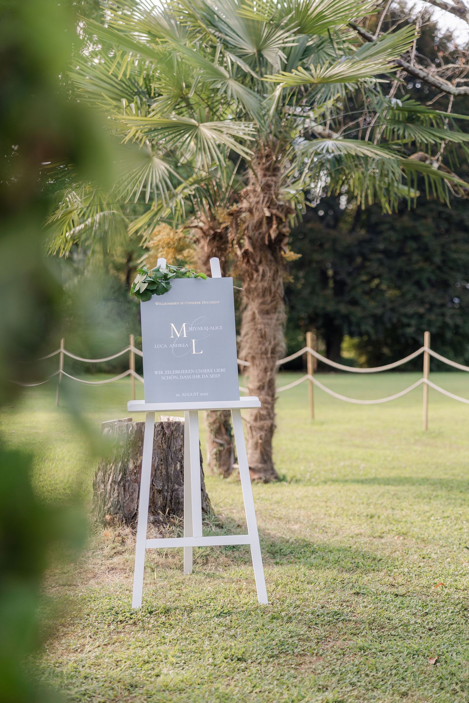 A white wooden easel with a gray sign stands on a grassy lawn with palm trees in the background.