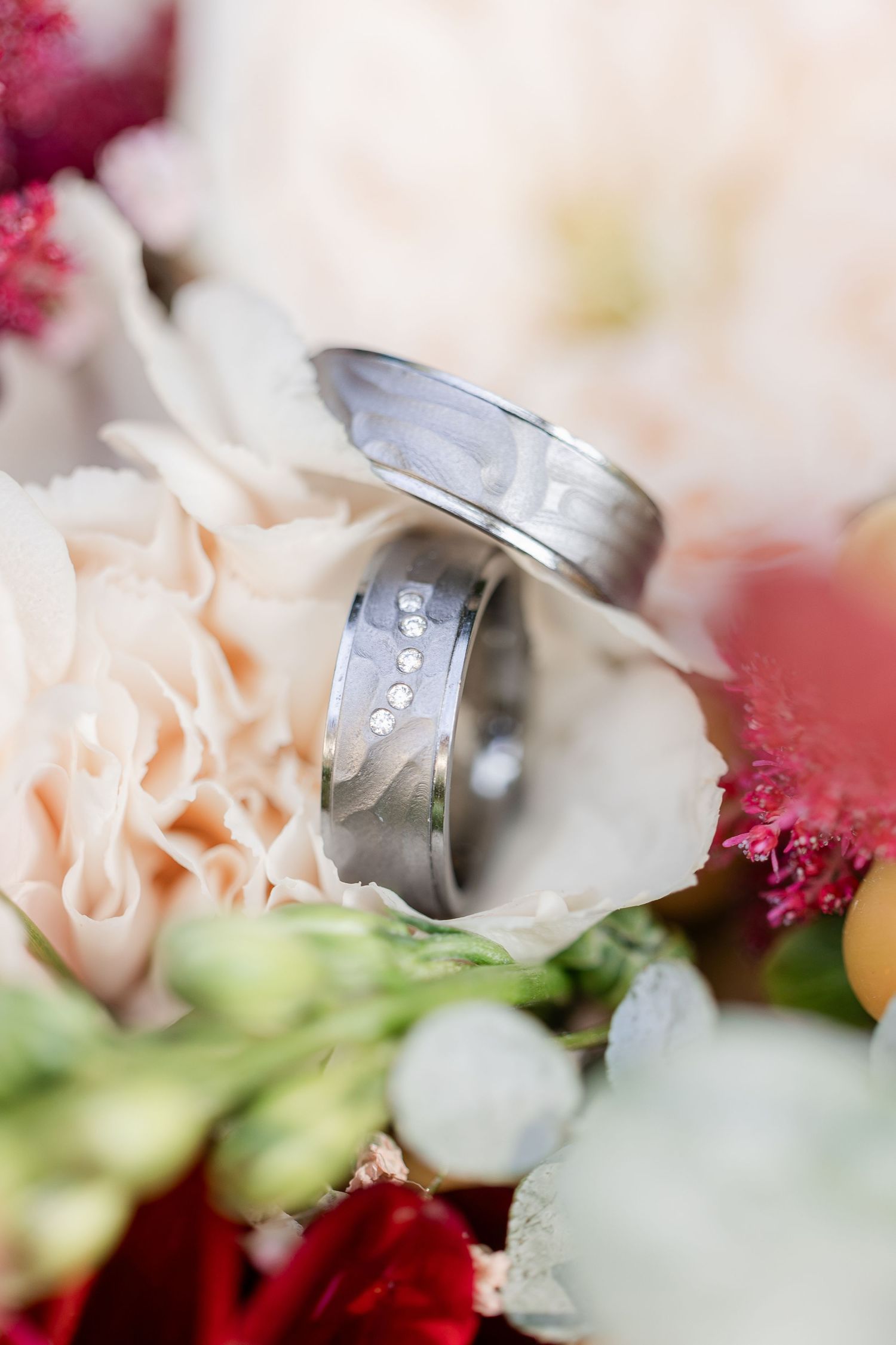 Close-up of two silver wedding bands nestled among pink and red flowers with soft blurred background.