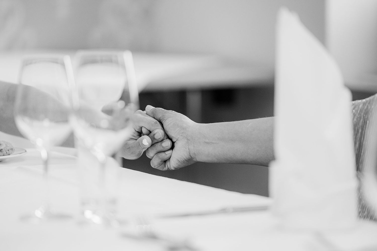 Black and white photo of two people holding hands at an elegantly set dining table with wine glasses.