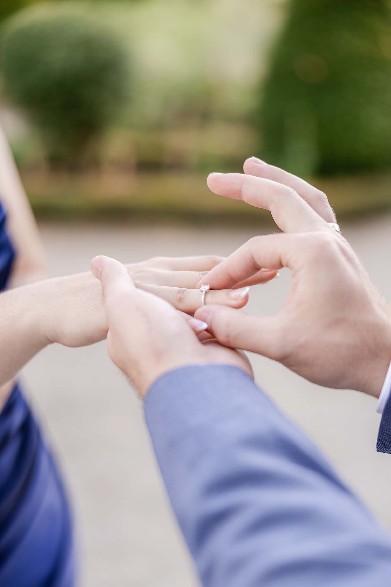 Person placing a diamond engagement ring on a woman's finger outdoors, with a blurred green garden background.