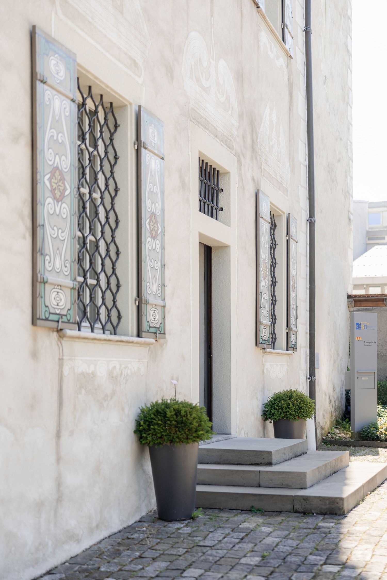 Small potted shrubs line the exterior wall of a white building with decorative window grates and stone steps.
