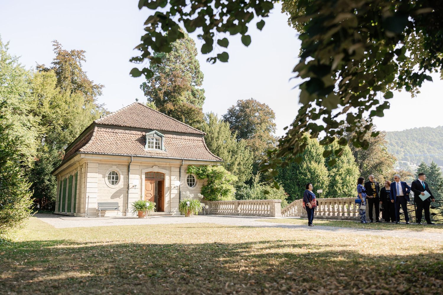 A small beige stone building with a hip roof sits among trees while guests gather outside on a sunny day.
