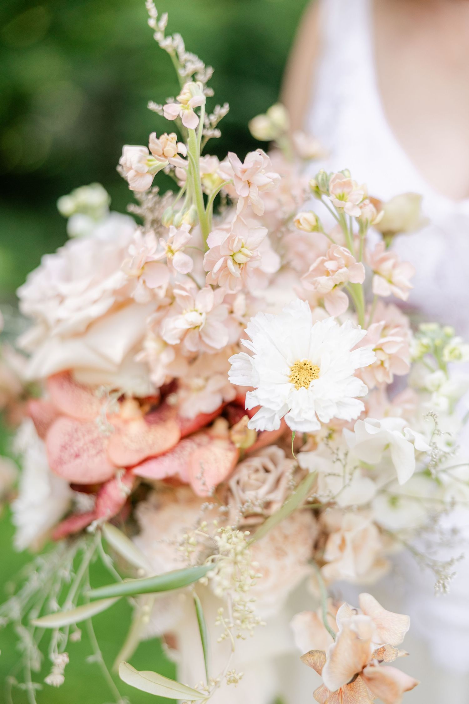 Bride holding a romantic bridal bouquet with white cosmos, blush roses, and delicate wildflowers against a green garden backdrop.