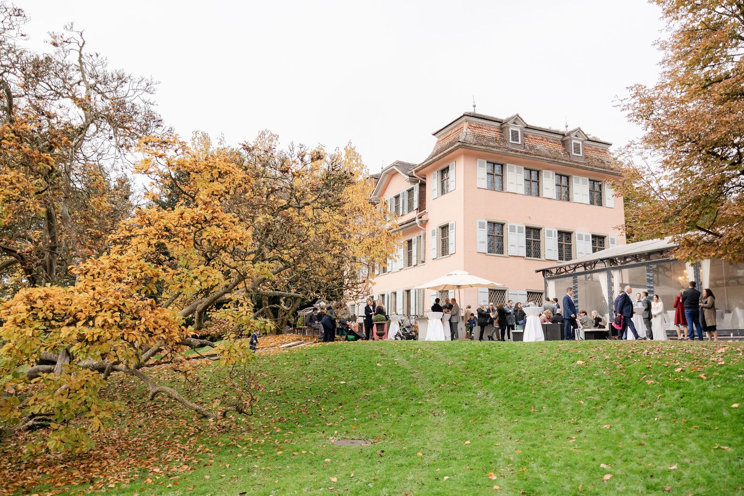 A large historic villa with pink facade surrounded by autumn trees and guests gathered on the lawn.