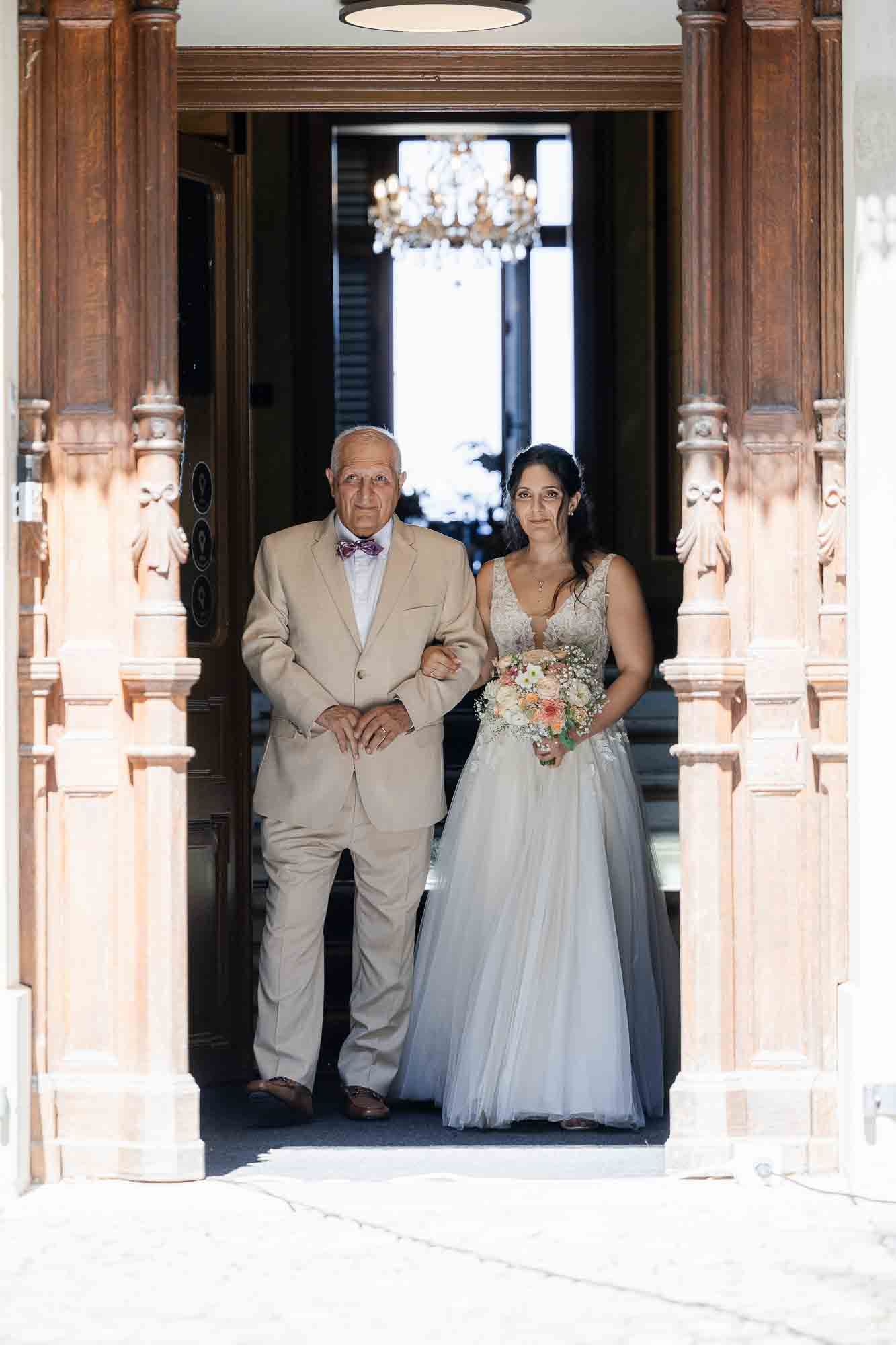 Bride in blue tulle gown holding bouquet walks with older man in tan suit through ornate wooden doorway.