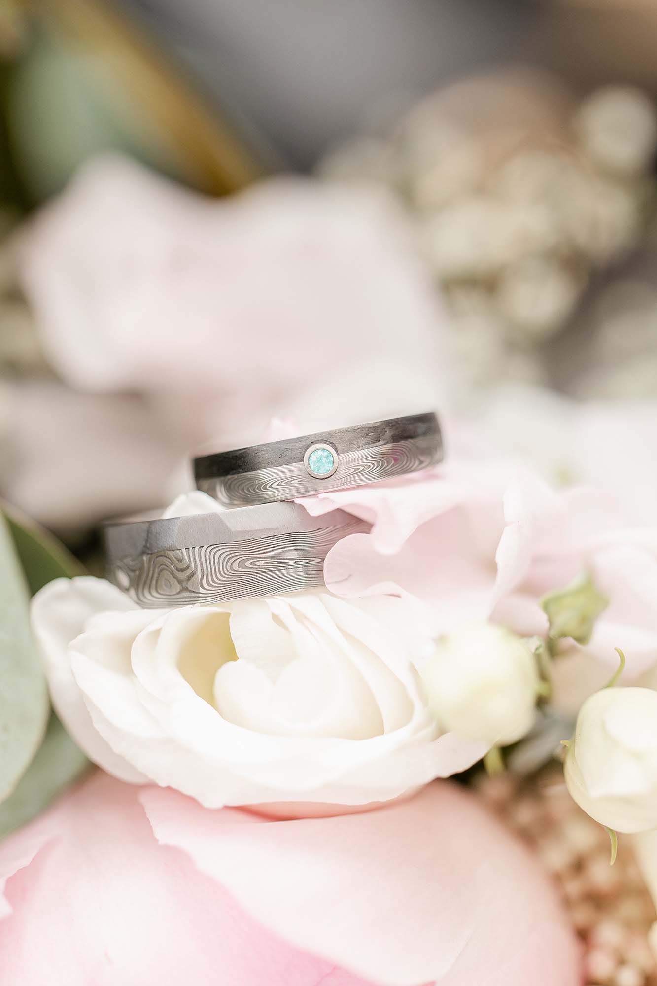 A silver damascus steel wedding ring with a small blue gemstone rests on delicate pink and white rose petals.