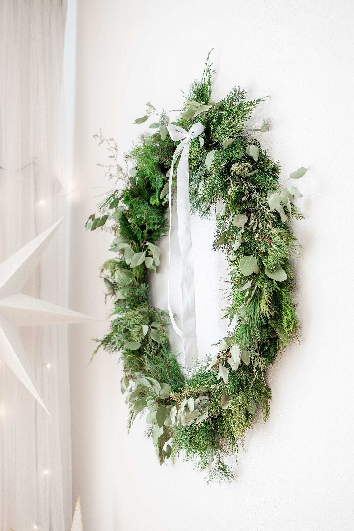 Fresh evergreen Christmas wreath with mixed greenery and white ribbon hanging against a light wall with twinkling lights.