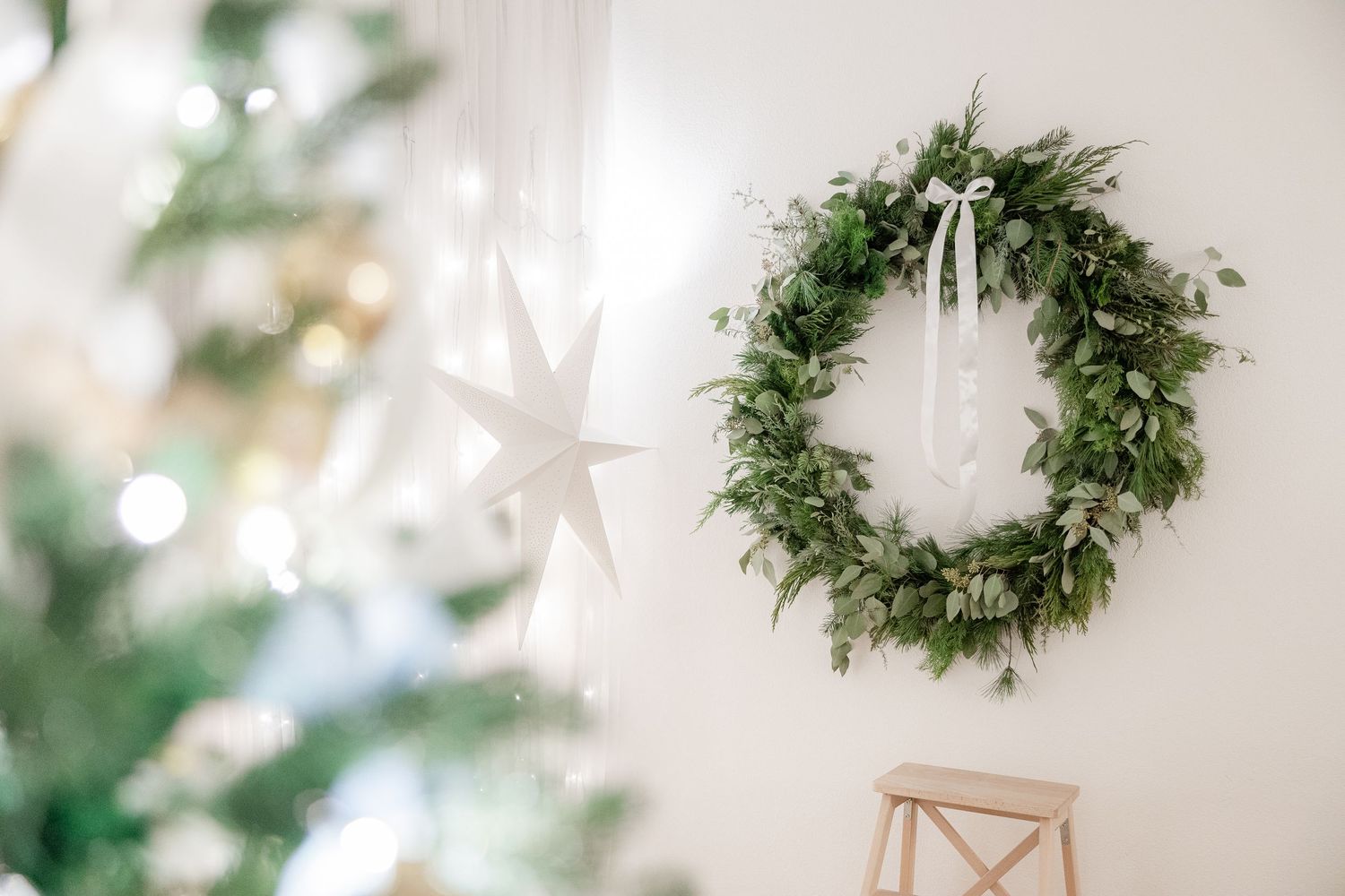 Fresh green holiday wreath hanging on white wall with white star decoration and blurred Christmas tree in background.