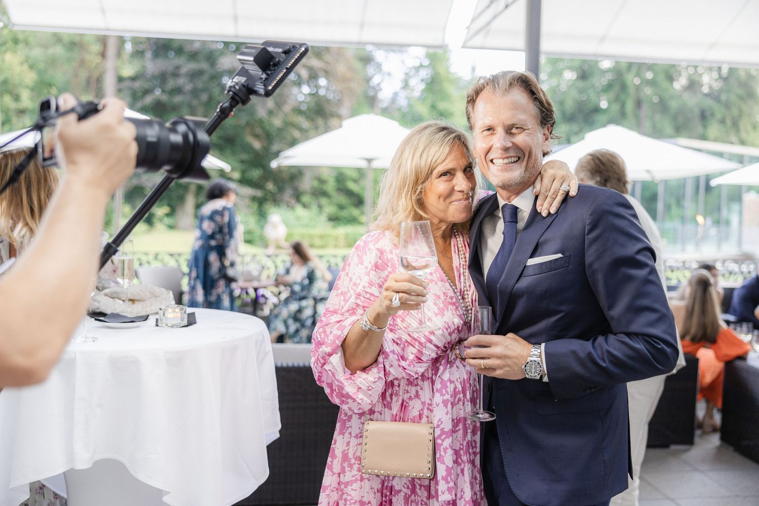 A couple poses together at an outdoor event with camera equipment and tables visible in the background.