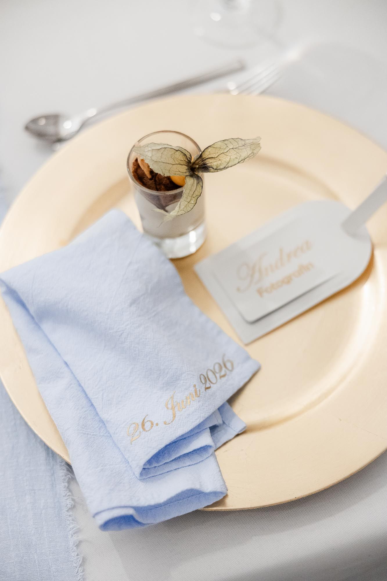 Elegant wedding table setting with gold charger plate, blue linen napkin dated 26 Juni 2026, place card, and decorative dessert cup.