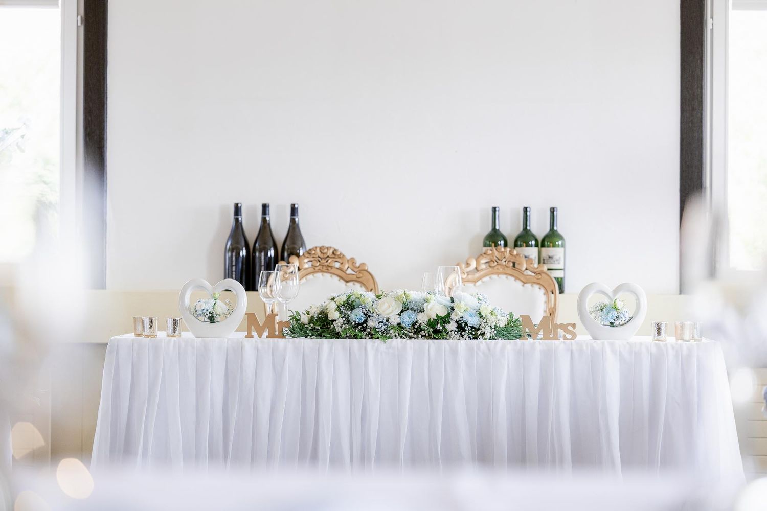 Elegant wedding sweetheart table with white tulle, floral garland, Mr. & Mrs. signs, and wine bottles.