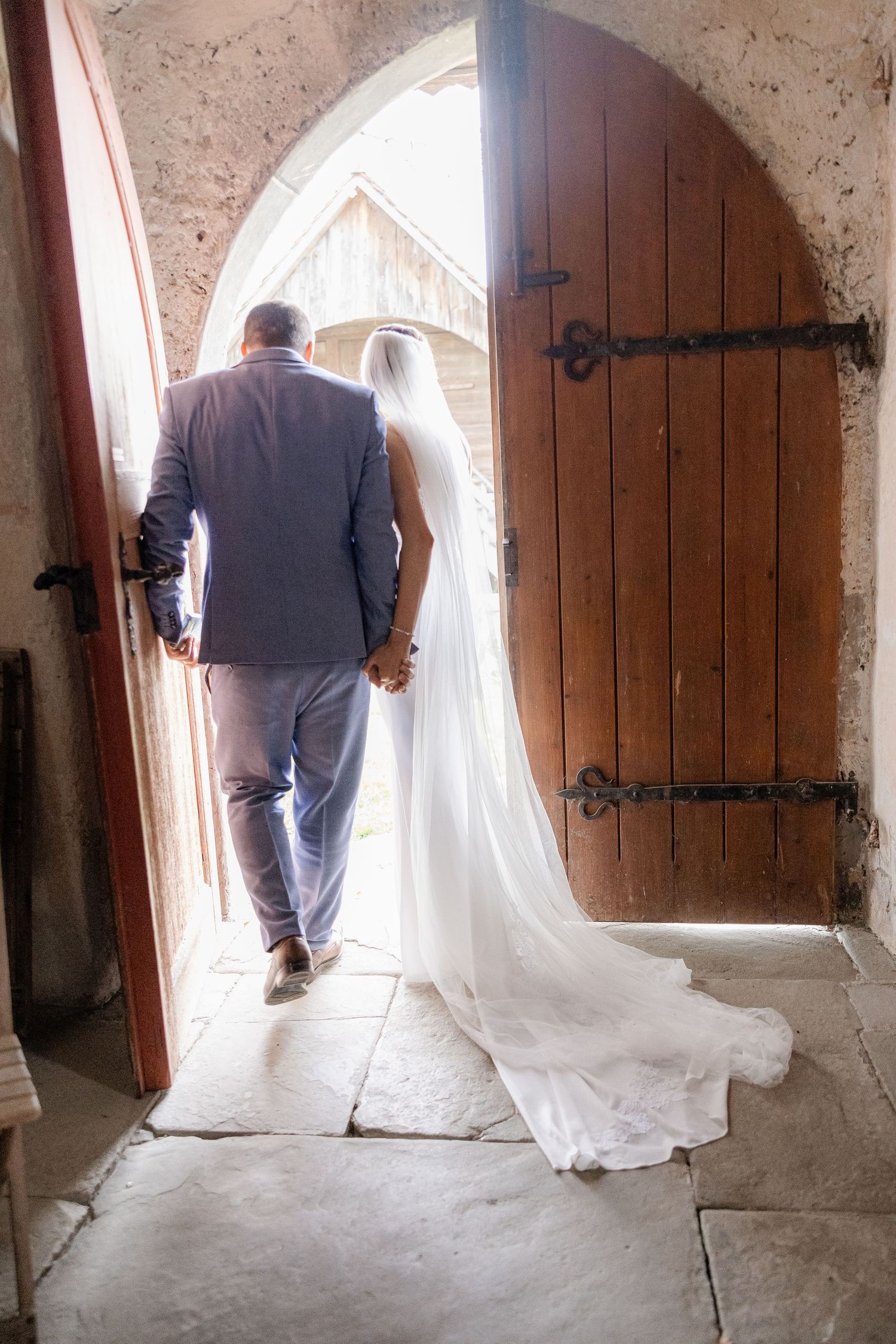 A couple in wedding attire walk through an arched stone doorway with a rustic wooden door at a church.