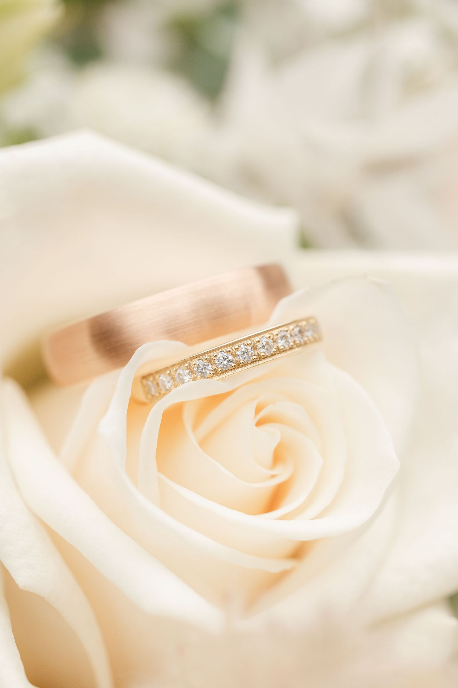 A delicate gold diamond wedding band rests atop a soft cream-colored rose in a romantic macro shot.