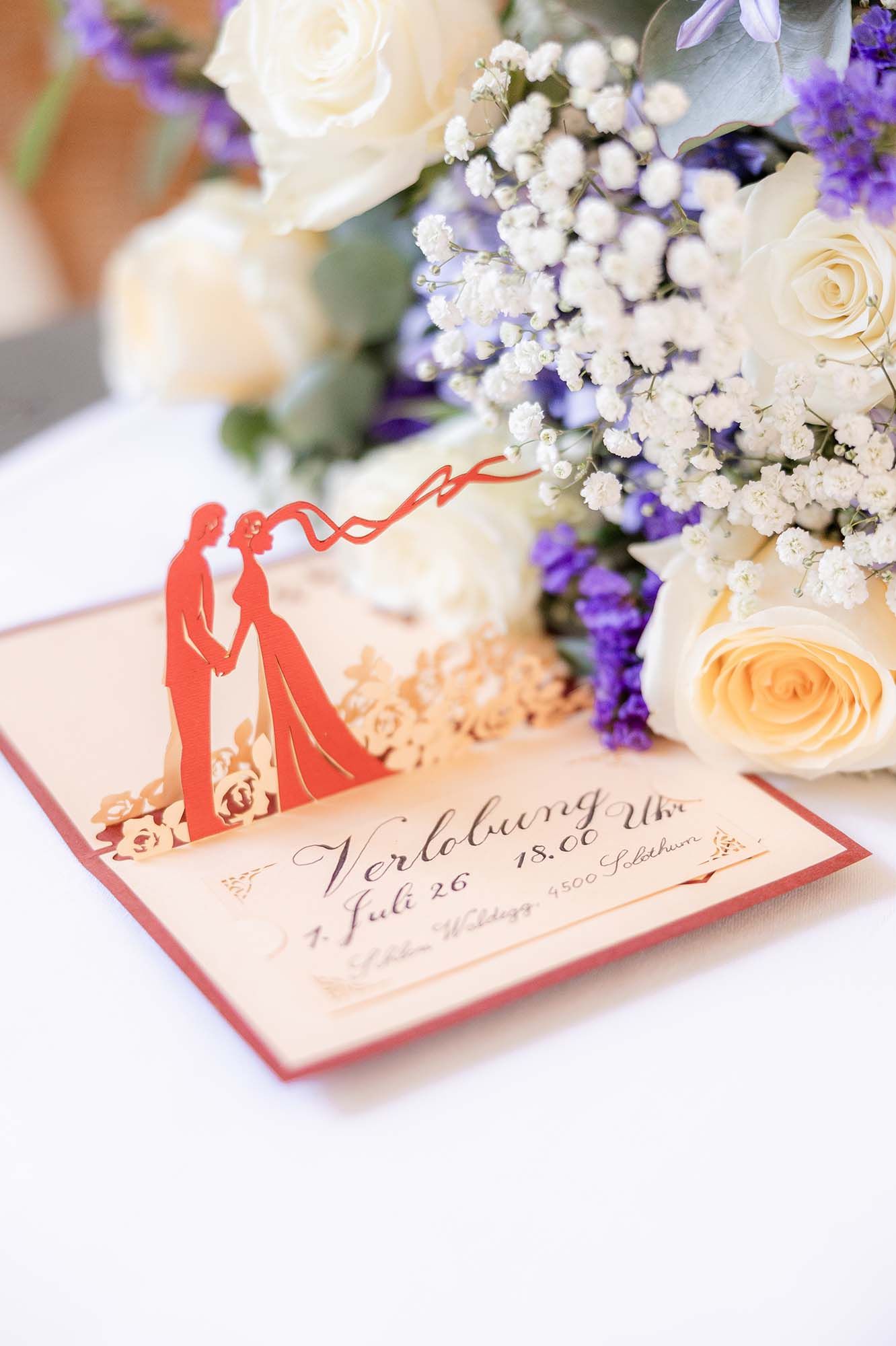 Red pop-up engagement card with bride and groom silhouette beside a bouquet of white roses and purple flowers.