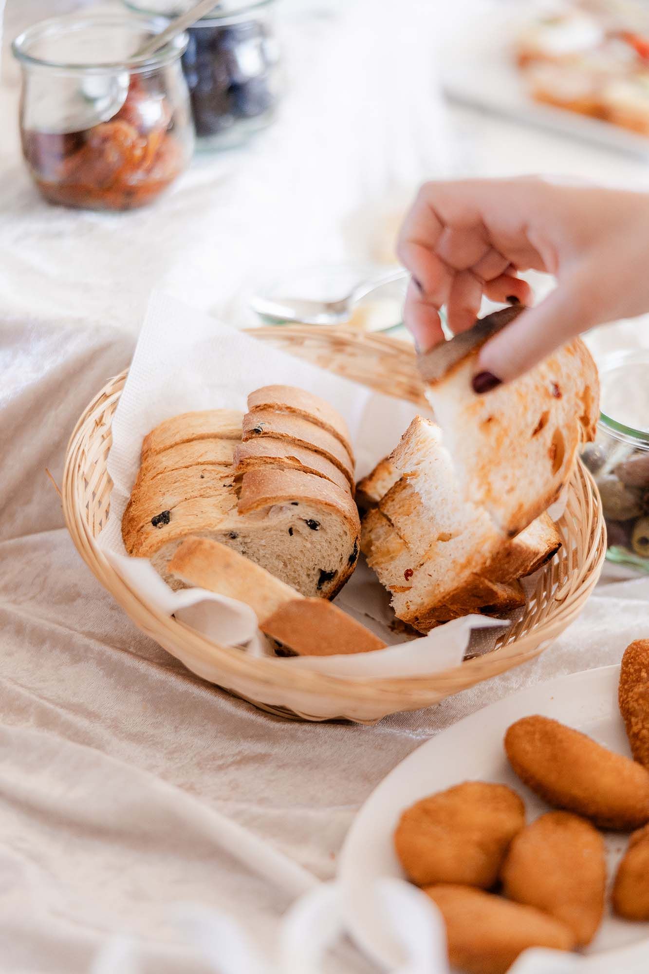 Hand reaching for sliced artisan bread with olives in a wicker basket lined with parchment paper on a white table.