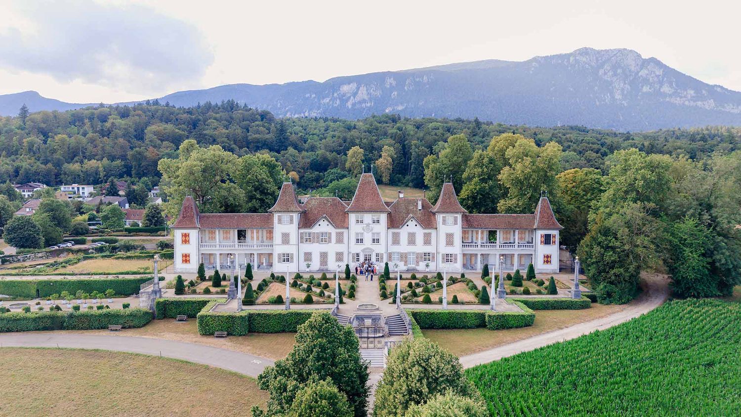 Aerial view of a grand French château with manicured gardens, surrounded by lush trees and mountains in the background.