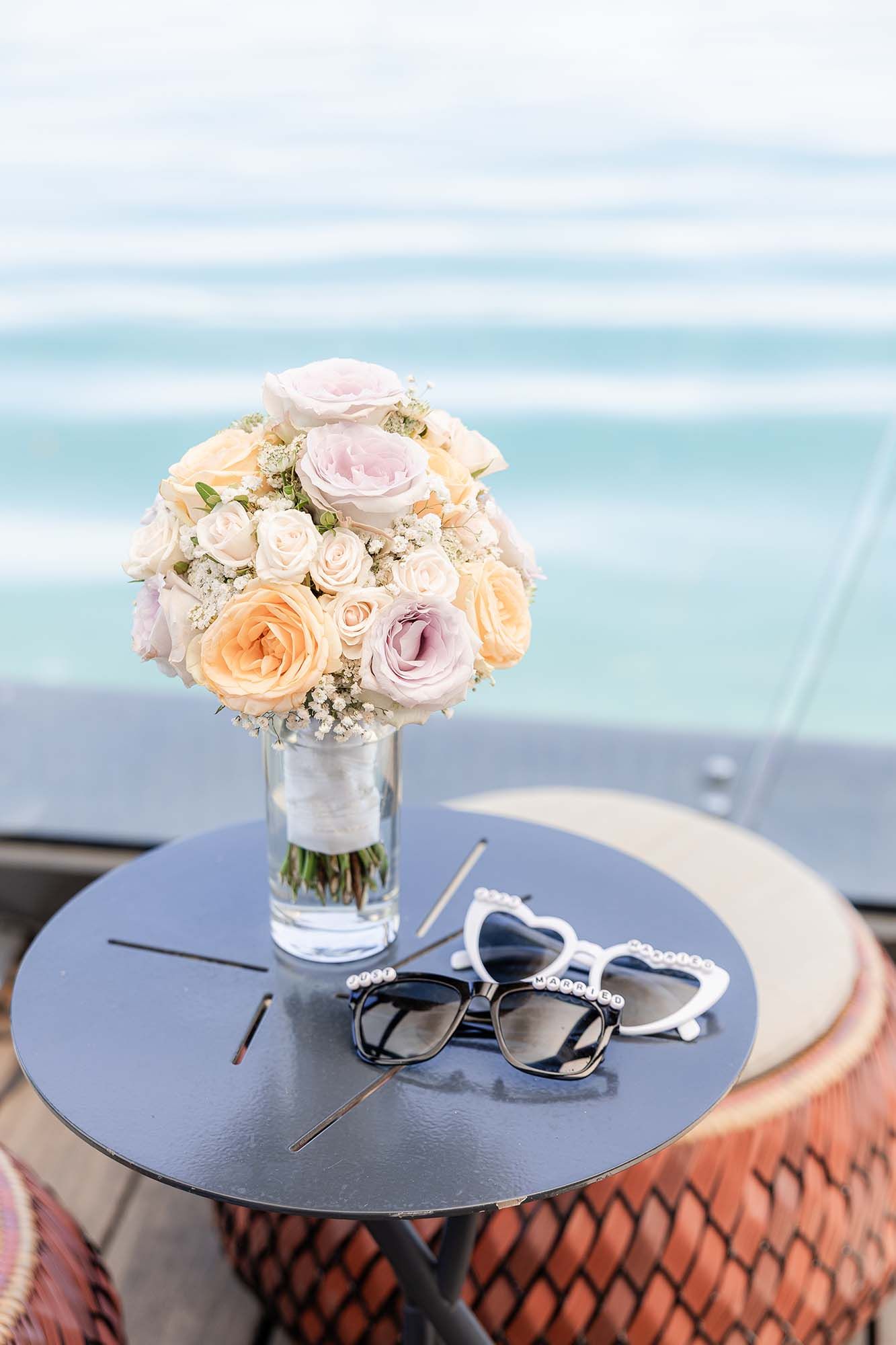 Elegant wedding bouquet of peach and pink roses in a vase beside two heart-shaped sunglasses on a table by the ocean.