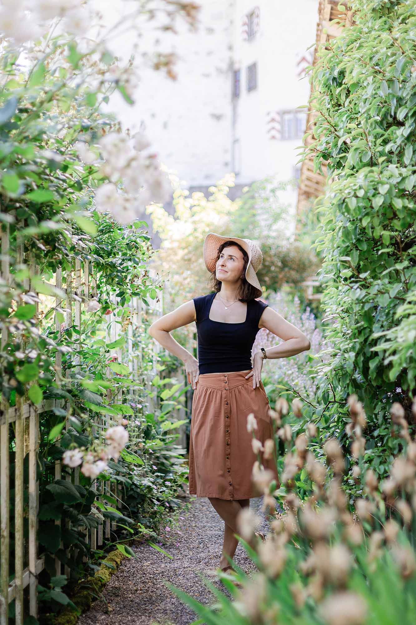 Woman in straw hat and brown skirt poses in a lush garden pathway surrounded by green plants.