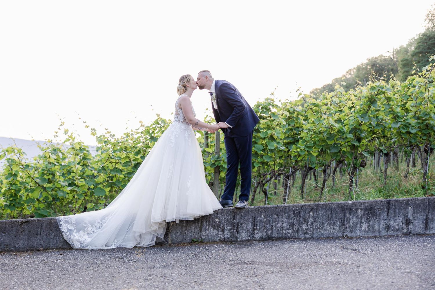Bride and groom share a romantic kiss at a vineyard wedding venue with lush green grapevines in the background.