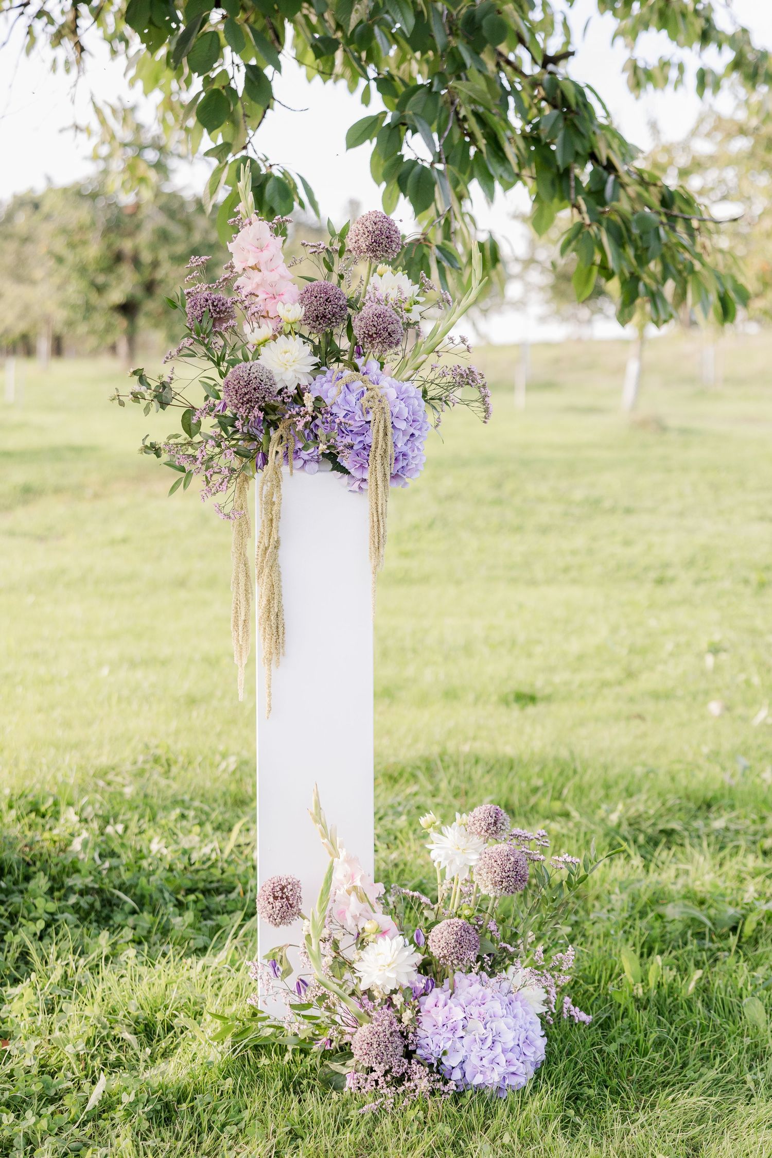 White decorative columns adorned with purple lilacs and wildflowers create a romantic garden display on lush grass.
