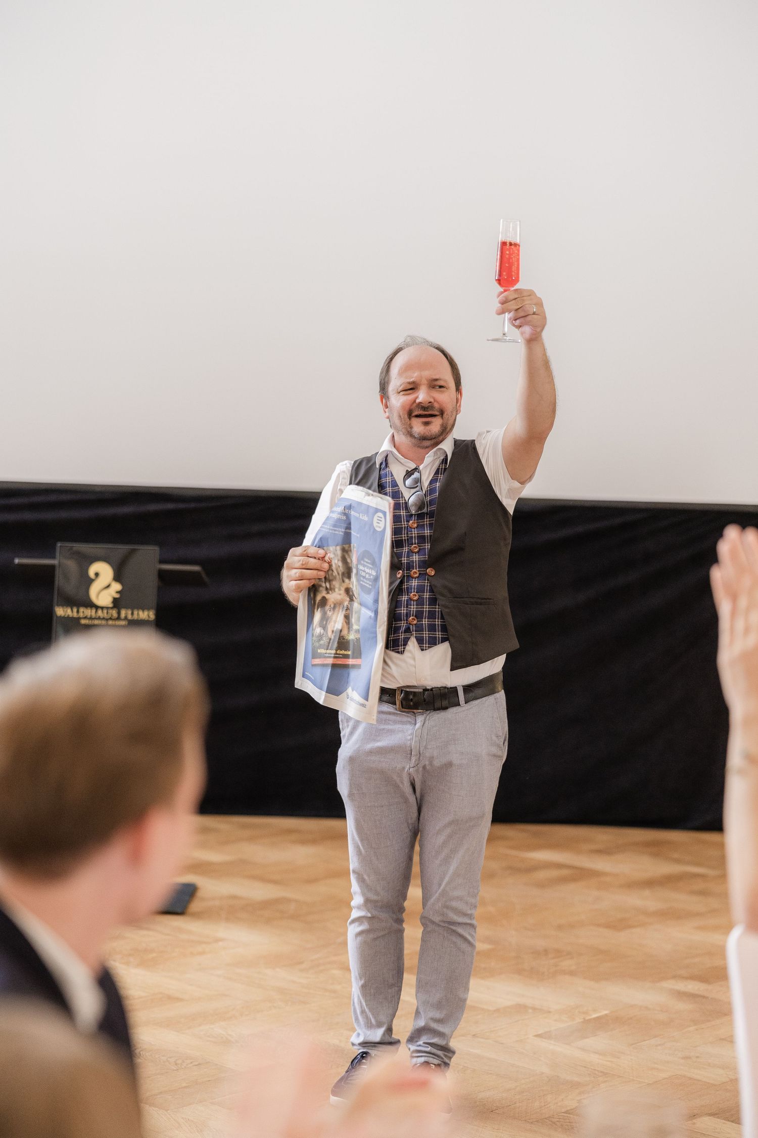 An enthusiastic person in a vest holds up items during what appears to be a presentation or performance.
