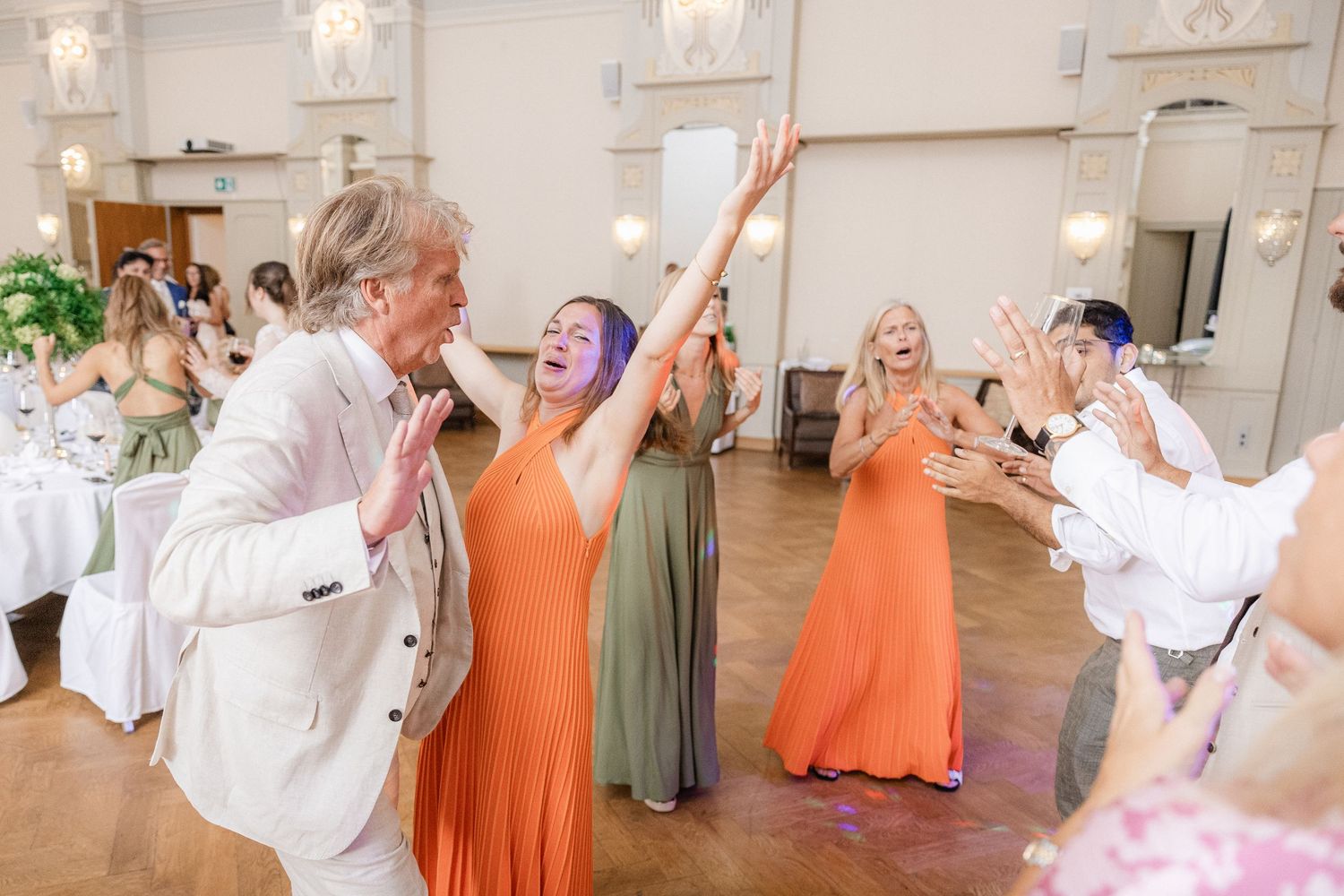 Wedding party in coordinated orange and white outfits dancing and celebrating together in an elegant indoor venue.