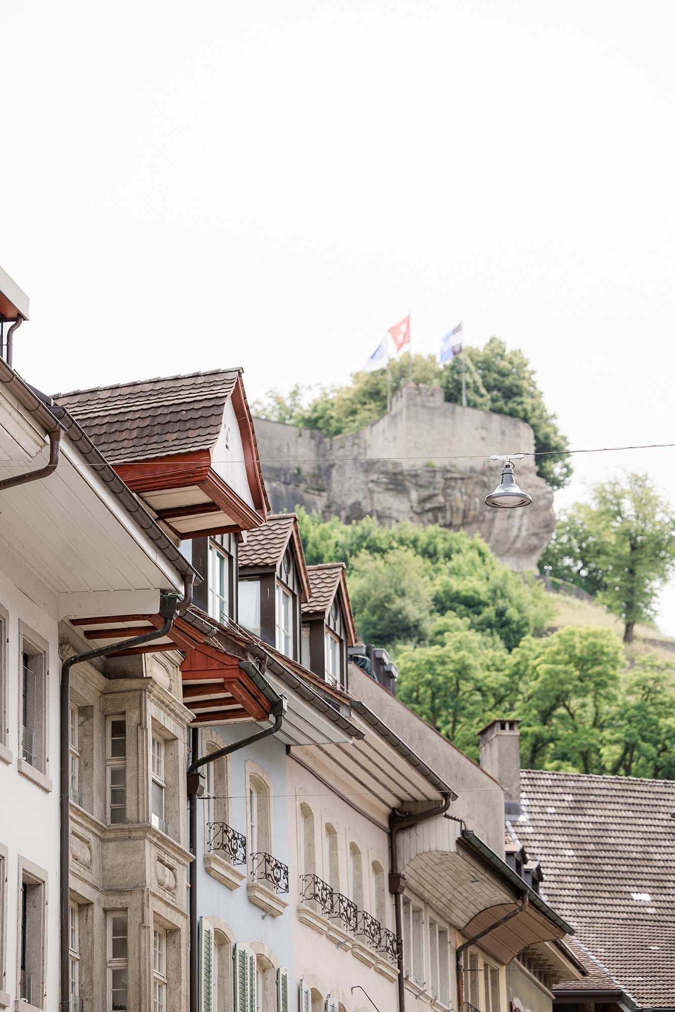 Historic European street with dormer-windowed buildings and a medieval castle on a rocky cliff with flags flying above.