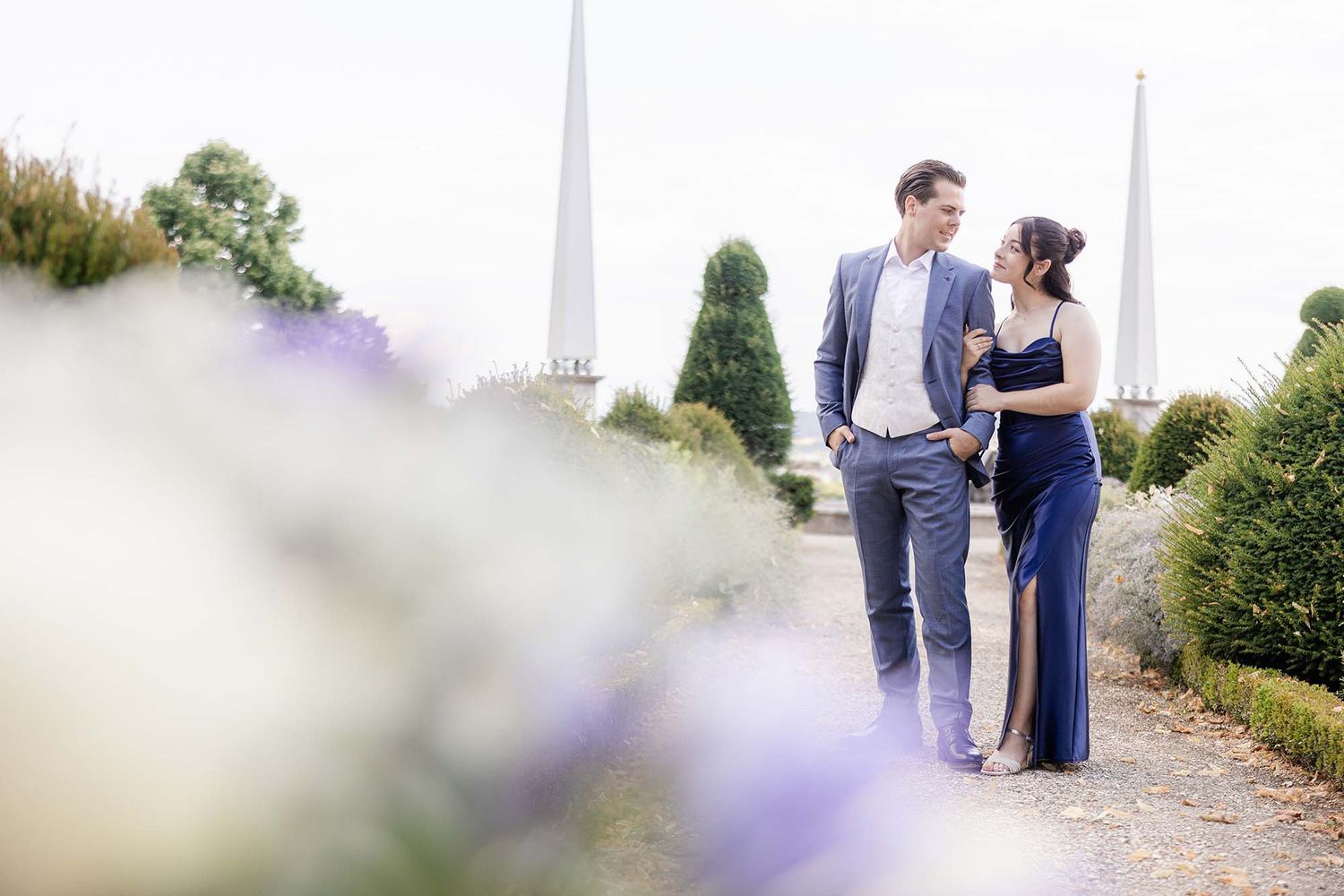 Couple in formal attire walking through a garden with purple flowers and tall hedges during an engagement photo session.