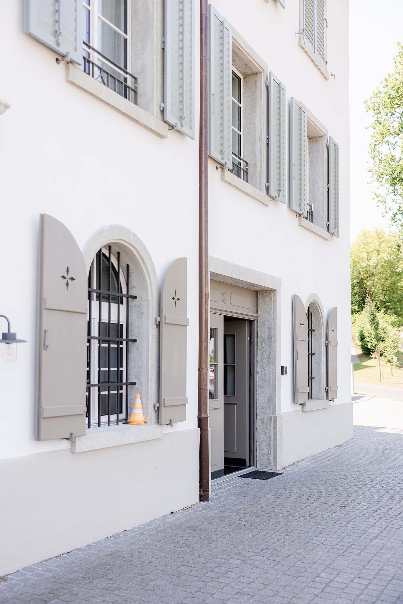 White European building facade with arched windows, gray shutters, iron grilles, and a wooden door on a cobblestone street.