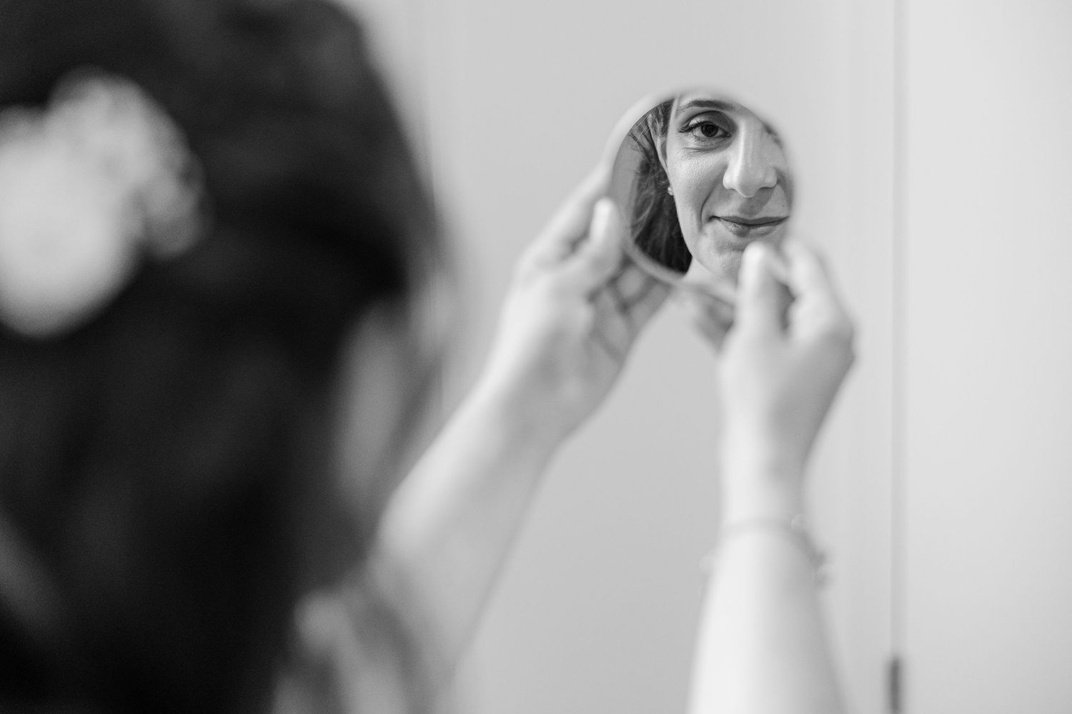 Black and white photo of a bride checking her makeup in a small handheld mirror before her wedding ceremony.