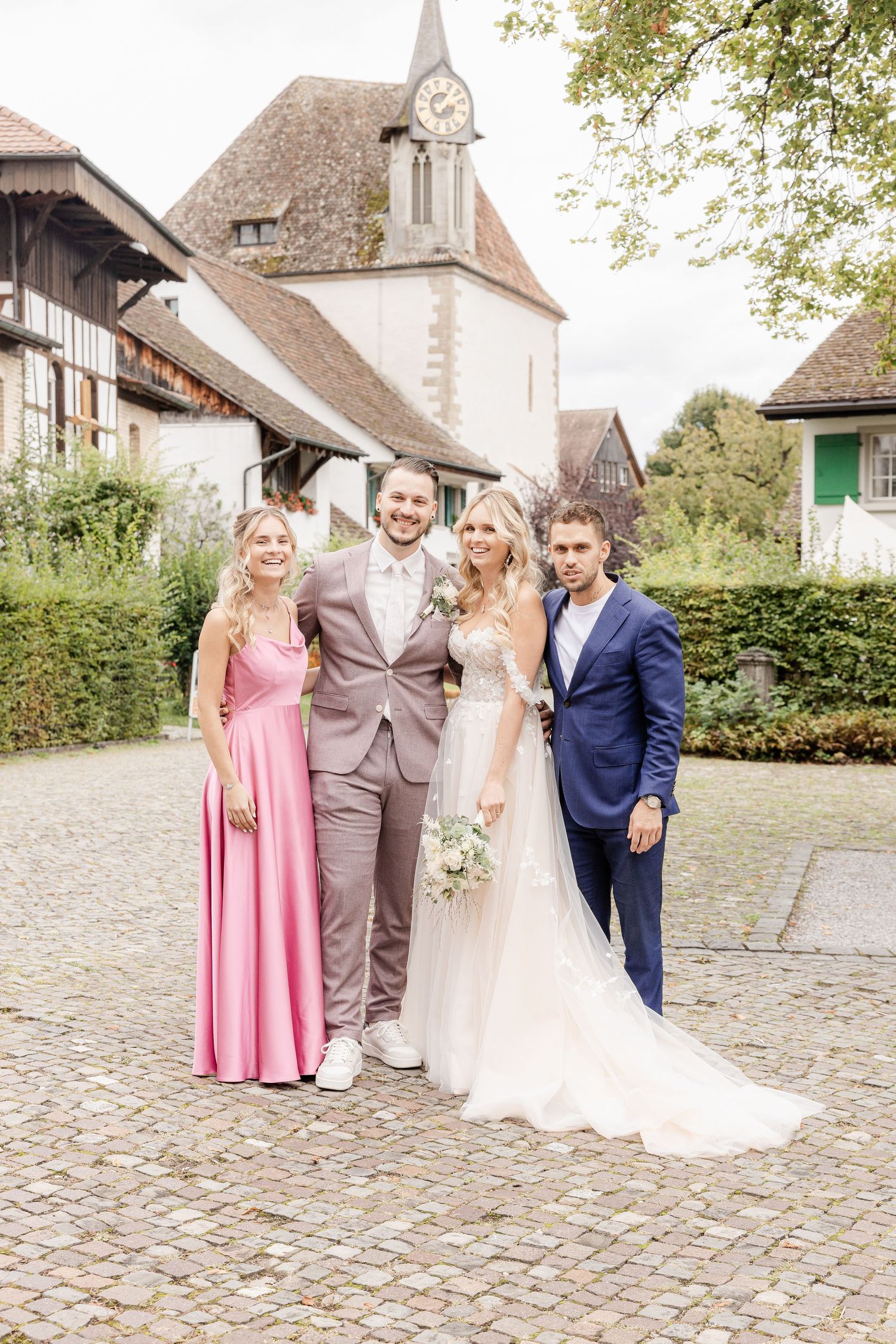 A wedding party poses on a cobblestone path in front of a historic church with timber-frame buildings in the background.