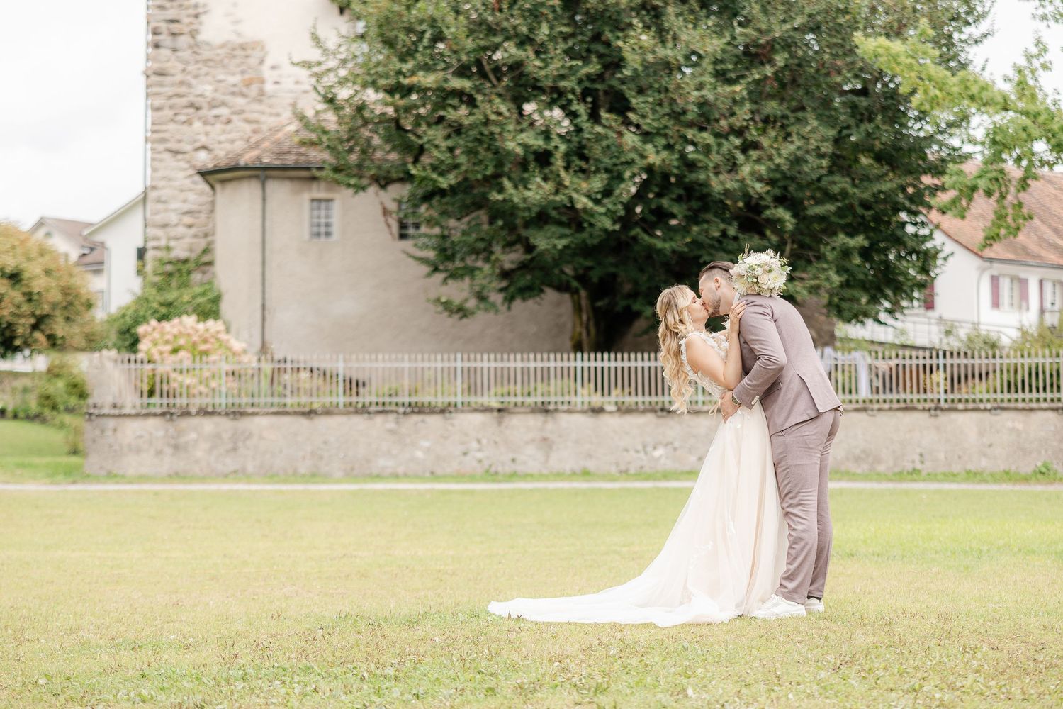 A newlywed couple embraces outdoors in a grassy field near stone walls and buildings on their wedding day.