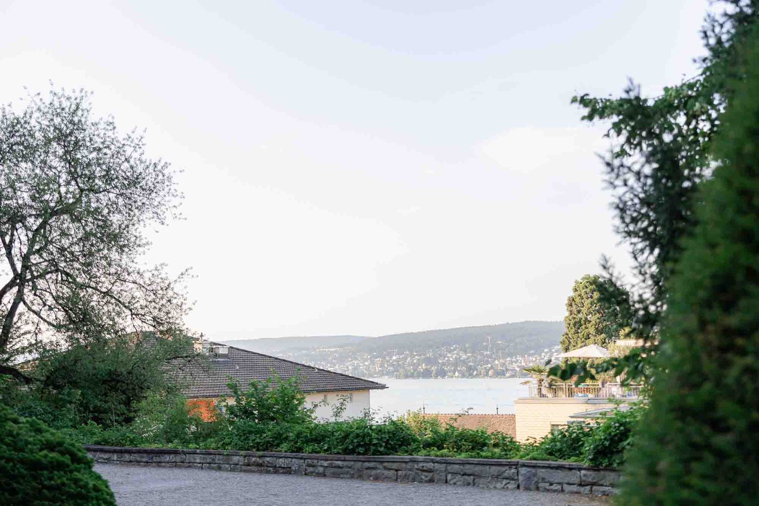 Scenic lakeside view with lush green trees, stone wall, and buildings overlooking calm water on an overcast day.