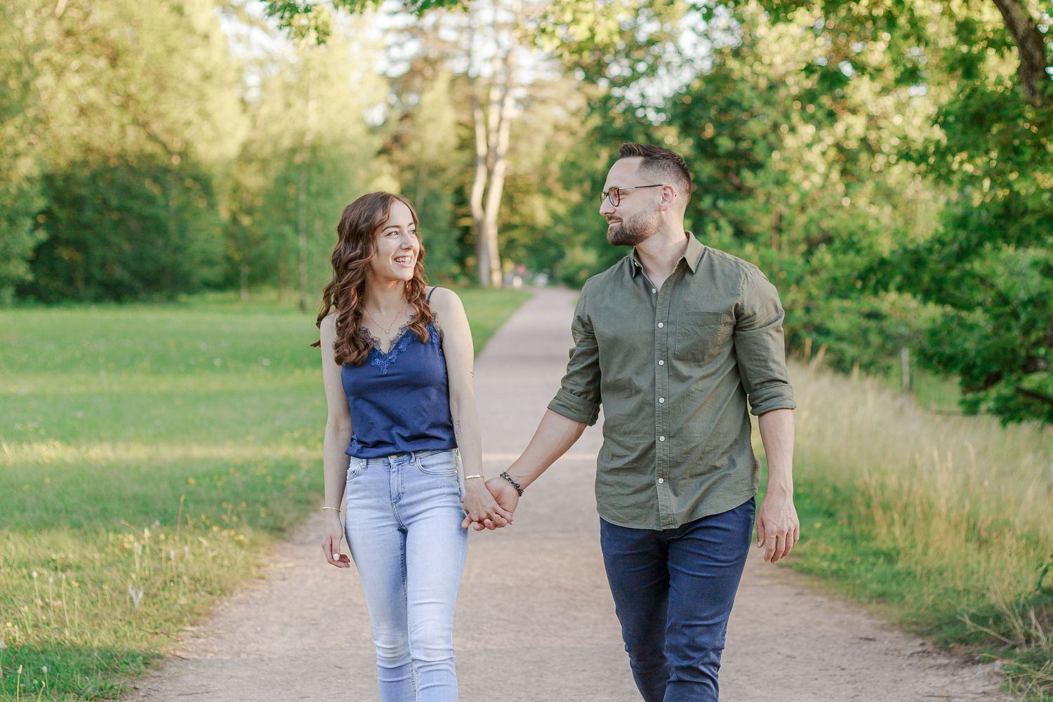 A couple walks hand in hand along a scenic path surrounded by lush greenery on a sunny day.