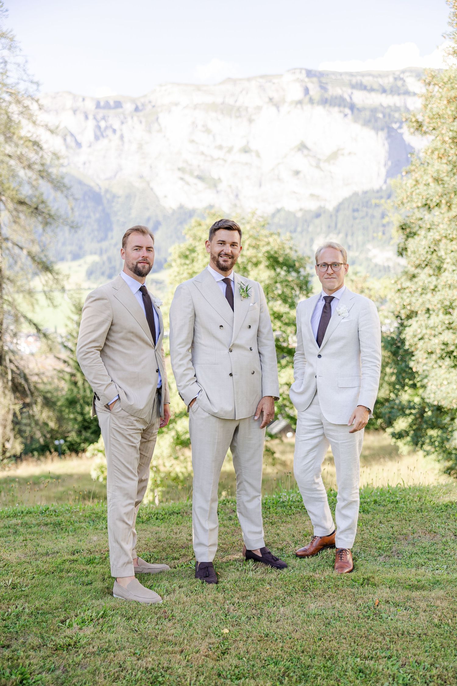 Groomsmen in light beige suits and colorful socks pose playfully on a grassy hillside with mountains in the background.