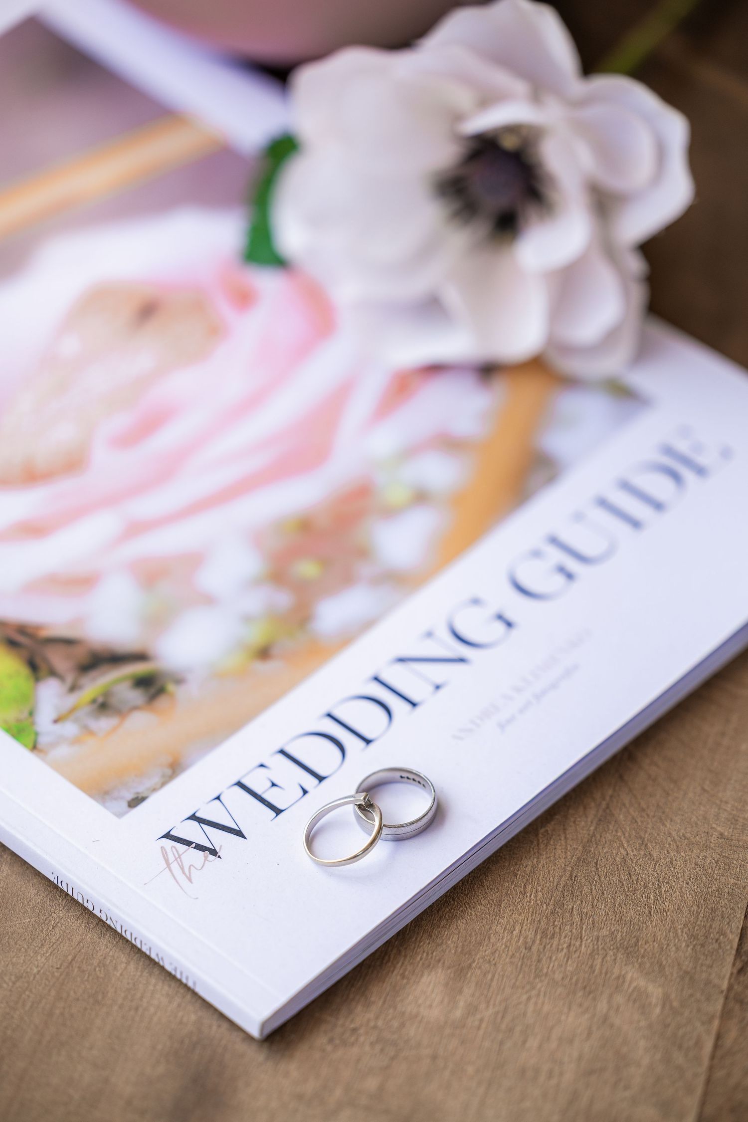 Two silver wedding rings rest on a Wedding Guide book with a white anemone flower in the background.