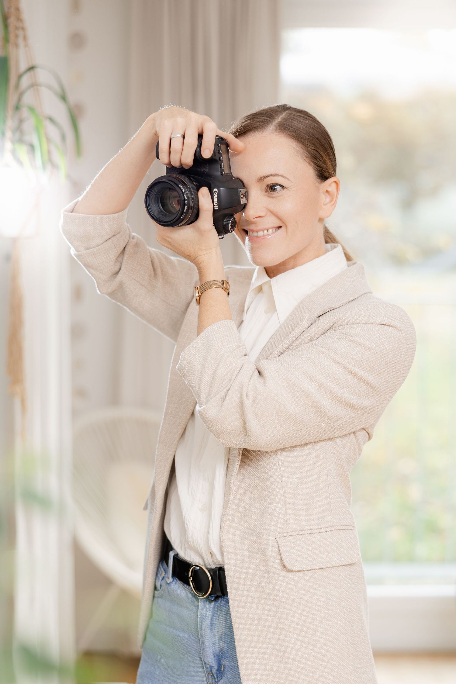 Photographer in beige blazer and jeans takes photos from different angles in bright interior space.